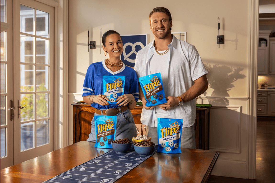 Professional football player and fashion designer, Kyle and Kristin Juszczyk, smile at the camera as they each hold a packet of Flipz chocolate-coated pretzels